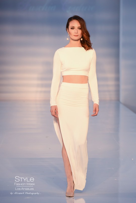 Image is of a femme person walking down a runway wearing a long-sleeved white two-piece dress with a long slit up one leg.