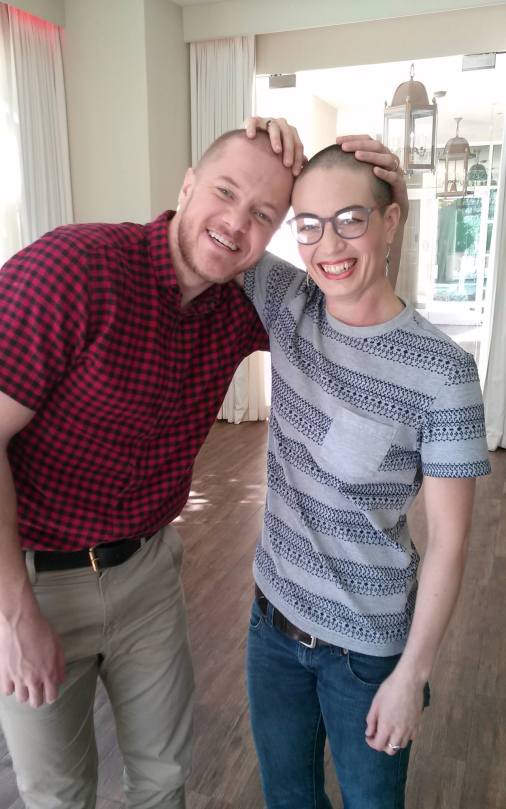 Dan Reynolds in red and black plaid shirt and khakis next to Charis Hill in a blue and grey top. Both have partially shaved heads and have their hands casually draped over the others' head.
