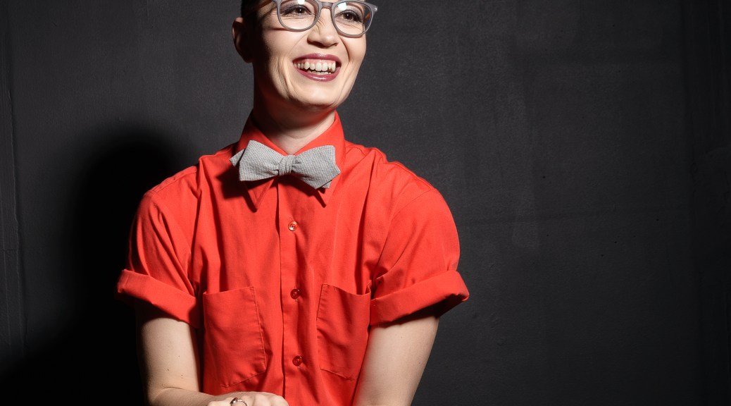 A light-skinned person sitting with right hand on right leg, wearing a red buttoned shirt, grey bowtie and glasses. They are in front of a dark grey background looking slightly away from the camera laughing.