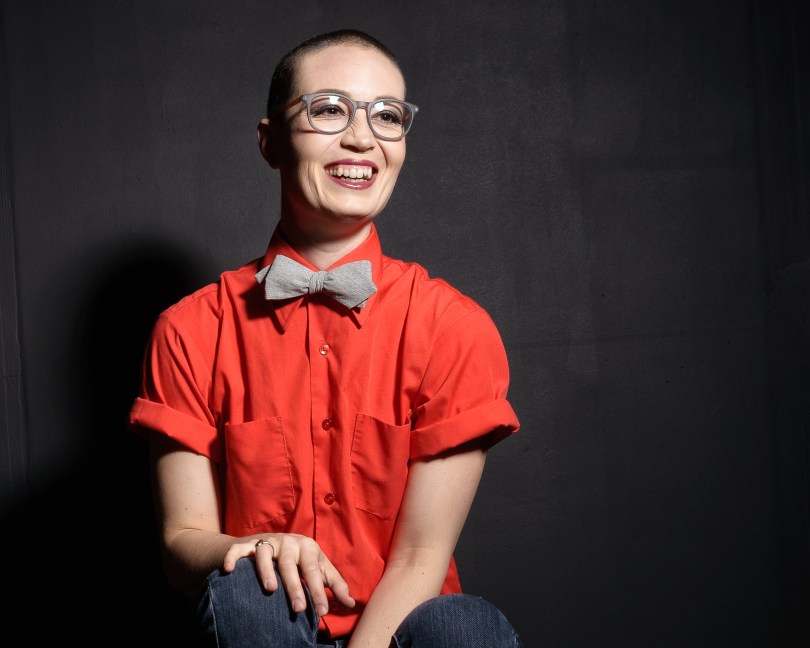A white person sitting with right hand on right leg, wearing a red buttoned shirt, grey bowtie and glasses. They are in front of a dark grey background looking slightly away from the camera laughing.