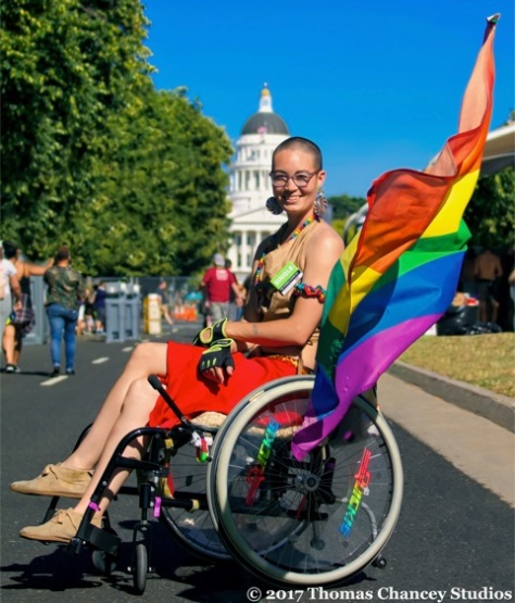 Image is of a light-skinned person outside in their wheelchair with a rainbow flag flying from it. The CA state capitol building is in the background.