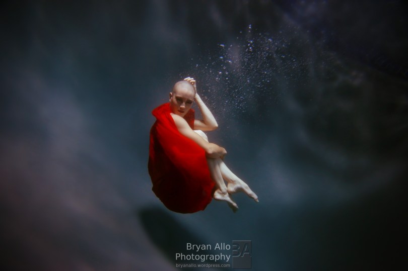 A picture from the side of a white bald person holding knees to chest in dark water, with red fabric surrounding their body, leaving their head, arms, and lower legs bare.