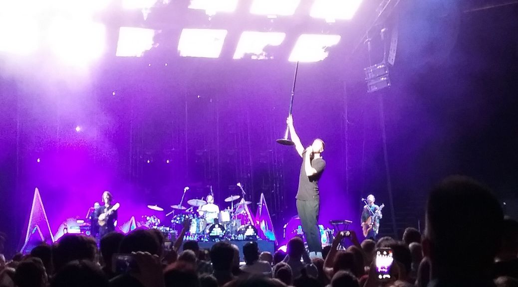 Dan Reynolds on stage holding a mic stand in the air. The rest of the band is visible in a purple haze