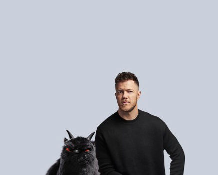 Dan Reynolds sits in front of a plan background with a black shirt on, looking at the camera. Next to him is a black beast with red eyes and two horns.