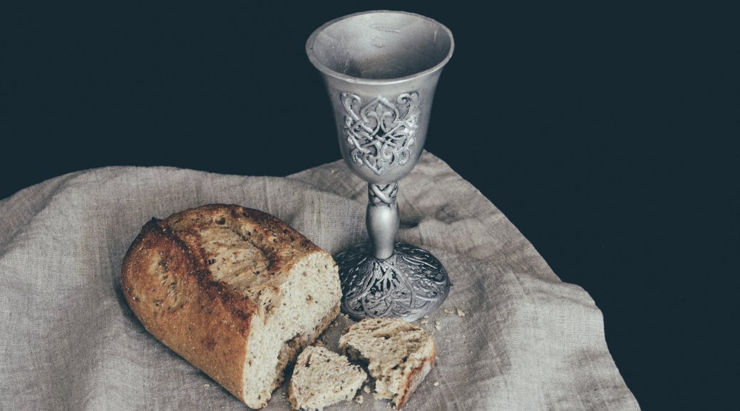 A chalice and half-loaf of bread resting on tan fabric with a black background