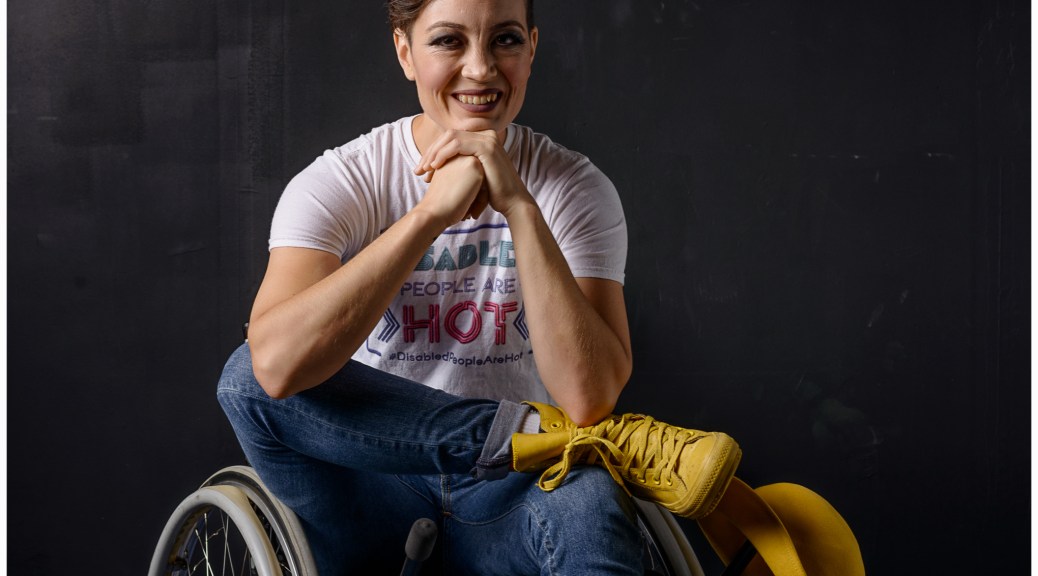 A studio portrait on black background of a white-appearing androgynous person in a wheelchair, one ankle crossed, resting on the opposite knee, and their hands under their chin as they smile at the camera