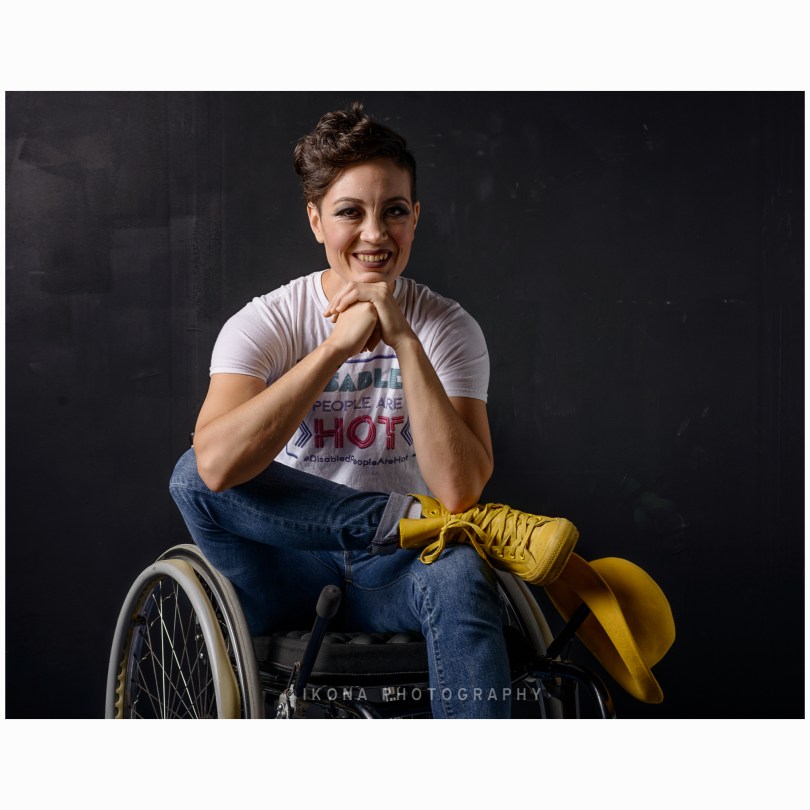 A studio portrait on black background of a white person in a manual wheelchair, one ankle crossed, resting on the opposite knee, and their hands under their chin as they smile at the camera