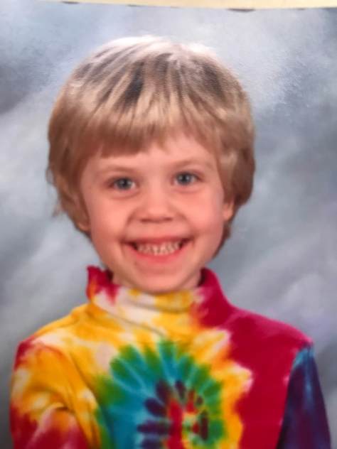 A white child with medium length blonde hair and a tie-dyed turtleneck shirt smiling in what appears to be a school portrait
