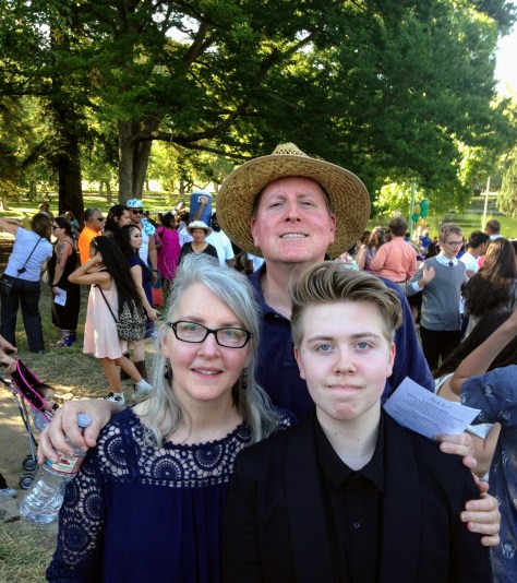 A family photo outside with a white mother, father, and son at his graduation from middle school. He wears a black robe. They all are smiling.