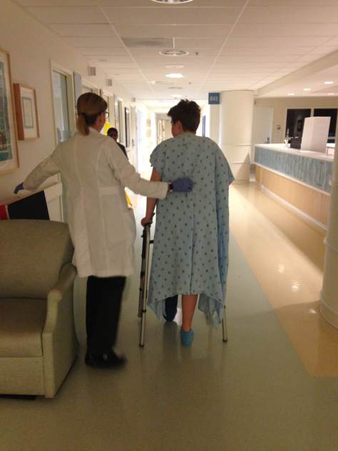 A picture from behind of a white-appearing healthcare provider with white lab coat with her hand slightly on the back of a young white teenager's back. He wears a blue hospital gown and takes a walk using a walker down a hospital hallway